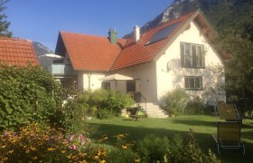 Ein Haus mit rotem Dach, umgeben von einem Garten mit Blumen und Liegestühlen, vor einer Bergkulisse.