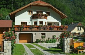Ein zweistöckiges Haus mit Holzbalkonen und Blumen, umgeben von grüner Landschaft.