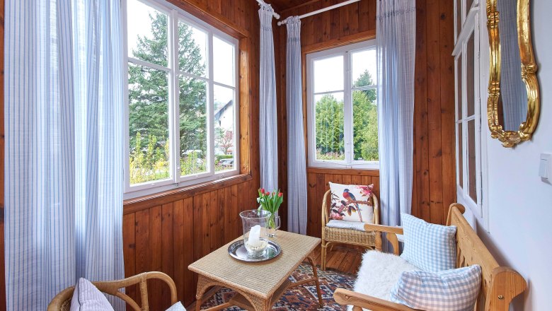 Top 1 - Wintergarten, © VP FeWo OG Ein gemütlicher Wintergarten mit Holzwänden, Korbmöbeln, blauen Vorhängen und einem Tisch mit Kerze und Blumen.