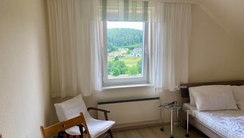 Helles Schlafzimmer mit Fensterblick auf grüne Landschaft, Bett, Sessel und Tisch.