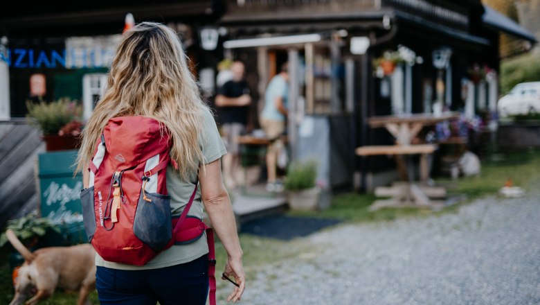 Frau mit rotem Rucksack und Hunde geht auf eine Berghütte zu.