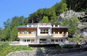 Ein modernes Geb&auml;ude mit Holzfassade vor einem bewaldeten Hang.