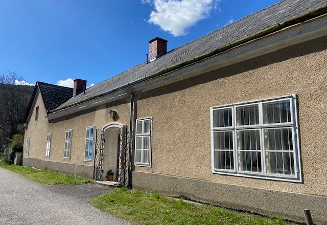 Langes, einstöckiges Gebäude mit mehreren Fenstern und einem Schrägdach unter blauem Himmel.