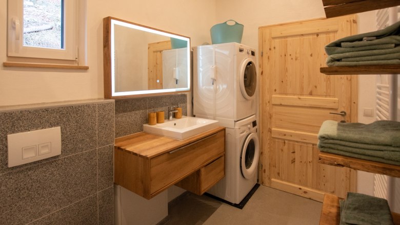 Modernes Badezimmer mit Holzwaschtisch, beleuchtetem Spiegel, Waschmaschine und Trockner. Handtücher auf Holzregalen.