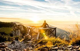 Person steht auf einer Holzplattform und blickt in den Sonnenuntergang über einer Berglandschaft.