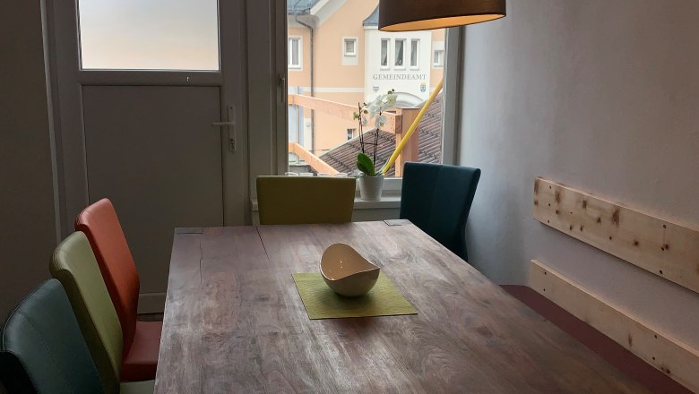 Essbereich mit Holztisch, bunten Stühlen und Hängelampe, Fenster mit Blick auf Gebäude.