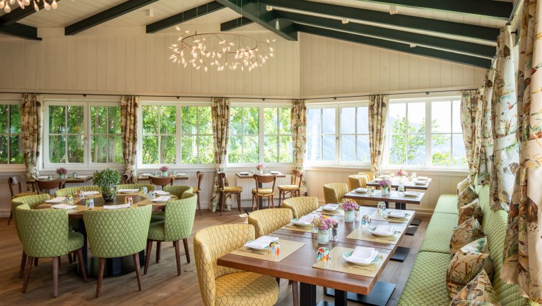 Ein heller Wintergarten mit gedeckten Tischen, grünen Stühlen und großen Fenstern.