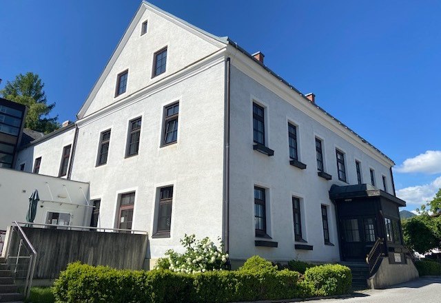 Ein großes, weißes Gebäude mit mehreren Fenstern und einem Giebeldach, umgeben von grünen Büschen und einem blauen Himmel im Hintergrund.