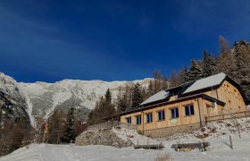 Ein Holzhaus im Schnee vor einer Bergkulisse unter klarem, blauem Himmel.
