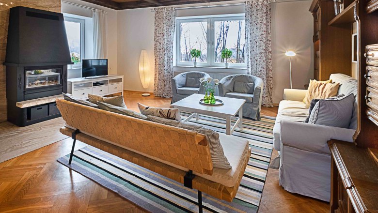Gemütliches Wohnzimmer mit Kamin, Sofa, Sessel und Fernseher. Holzboden und Decke, gestreifter Teppich, große Fenster mit Vorhängen.