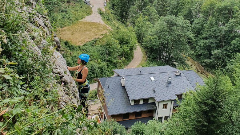 Person klettert an einer Felswand mit Helm und Kletterausrüstung, Gebäude und Wald im Hintergrund.