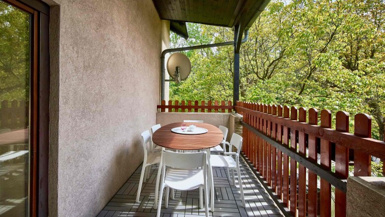 Ein Balkon mit Holztisch, weißen Stühlen und Blick auf grüne Bäume.