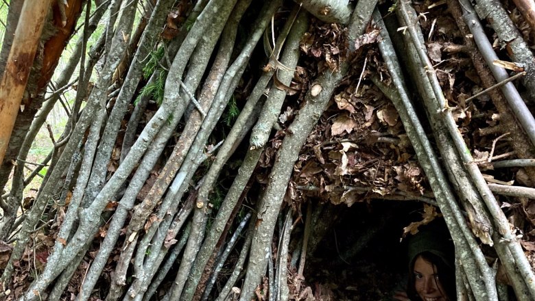 Eine Person schaut aus einem selbstgebauten Unterschlupf aus Ästen und Blättern im Wald hervor.