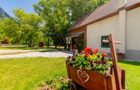 Ein blumengeschmückter Holzwagen vor einem Gebäude mit grünem Fensterrahmen, umgeben von Bäumen und Wiese.