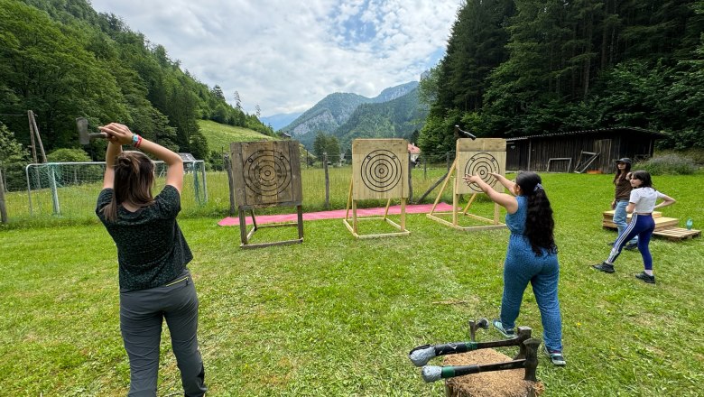 Personen werfen Äxte auf Zielscheiben im Freien, umgeben von Wald und Bergen.