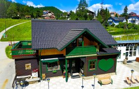 Ein traditionelles Holzhaus mit grünem Herzmotiv und Terrasse im Sommer, umgeben von grünen Hügeln und blauem Himmel.