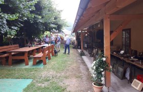Ein Gastgarten mit Holztischen und Bänken, Menschen im Gespräch, ein Hund, und ein überdachter Bereich mit Flohmarkt.