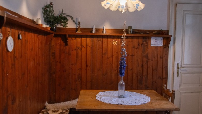 Rustikaler Raum mit Holzwänden, Tisch und Bank. Vase mit blauen Blumen auf Tischdecke. Lampe mit Blumenmuster an der Decke.
