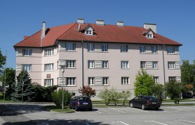 Wohngeb&auml;ude mit rotem Dach und mehreren Fenstern, umgeben von B&auml;umen und geparkten Autos.