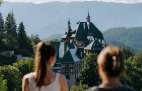 Zwei Menschen, mit dem Rücken zur Kamera, blicken auf eine malerische Berglandschaft. Im Vordergrund sieht man das Südbahnhotel, eingebettet in üppiges Grün. Im Hintergrund erheben sich Berge unter einem klaren blauen Himmel.