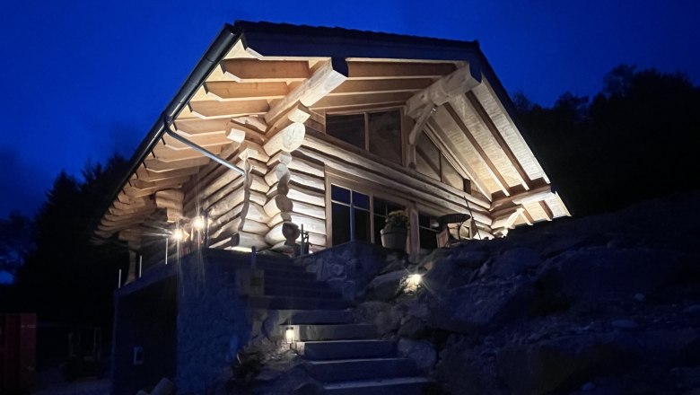 Ein beleuchtetes Holzchalet bei Nacht mit steinernen Treppenstufen im Vordergrund.