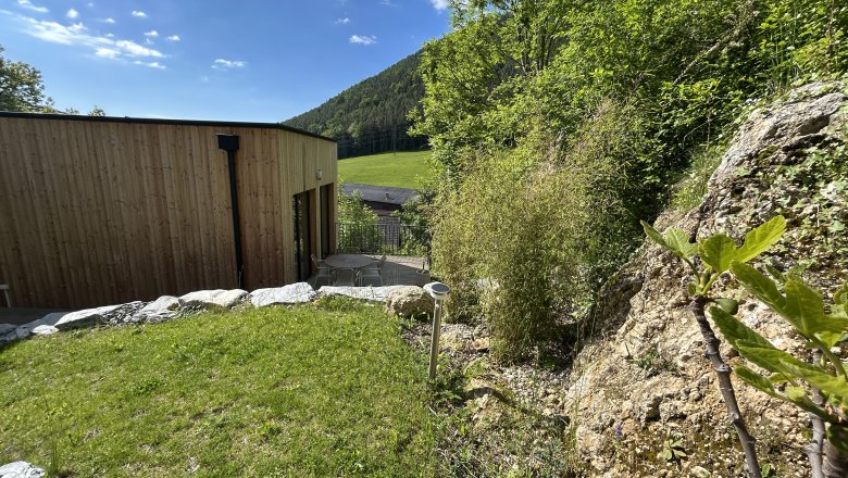 Ein modernes Tiny House mit Holzverkleidung steht in einer gr&uuml;nen, h&uuml;geligen Landschaft. Im Vordergrund sind Felsen und Pflanzen zu sehen.