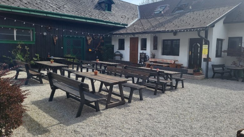 Ein Gastgarten mit Holzbänken und Tischen auf Kiesboden vor einem traditionellen Gebäude.