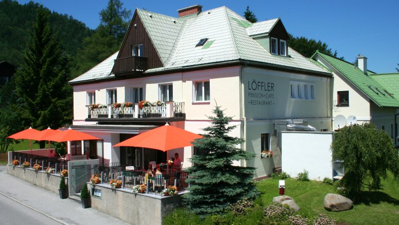 Ein zweistöckiges Gebäude mit der Aufschrift 'Löffler Pension-Café-Restaurant', umgeben von Bäumen und Blumen, mit orangenen Sonnenschirmen auf der Terrasse.