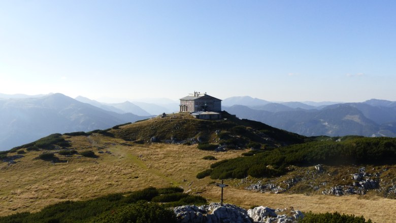 Berghütte auf einem Hügel mit Bergpanorama im Hintergrund.