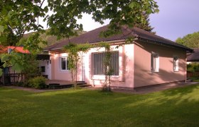 Ein rosa gestrichenes Ferienhaus mit dunklem Dach, umgeben von einem gr&uuml;nen Garten und B&auml;umen.