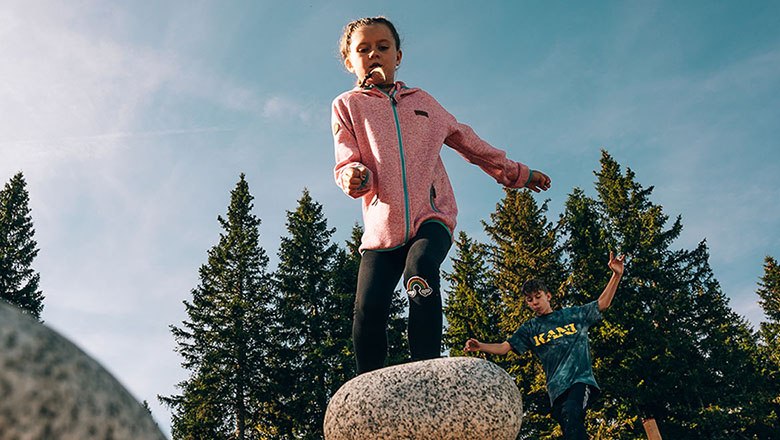 Kinder spielen auf Wackelsteinen vor einem Hintergrund aus B&auml;umen und blauem Himmel.