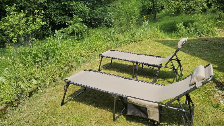 Zwei Liegestühle auf einer Wiese mit Blick auf bewaldete Berge unter blauem Himmel.