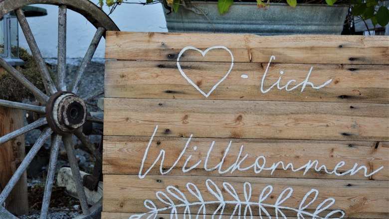 Holzschild mit der Aufschrift 'Herzlich Willkommen' und einem Herzsymbol, daneben ein altes Wagenrad.