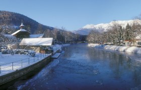 Winterlandschaft in Payerbach entlang der Schwarza mit schneebedeckten B&auml;umen und Bergen im Hintergrund.