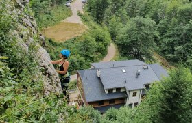 Person klettert an einer Felswand mit Helm und Kletterausrüstung, Gebäude und Wald im Hintergrund.