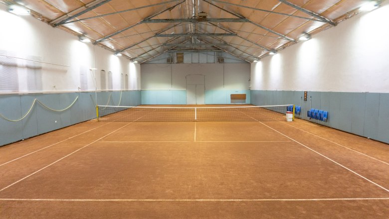Innenansicht eines leeren Tennisplatzes in einer Halle mit braunem Bodenbelag und blauen W&auml;nden.