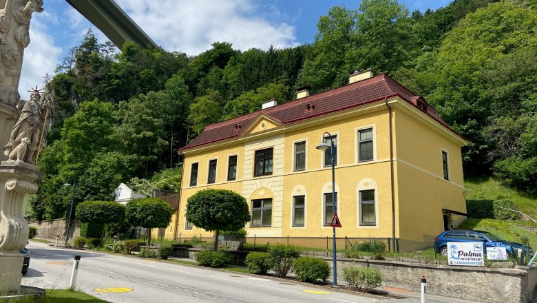 Apartment Palma, © Wiener Alpen Gelbes Gebäude mit rotem Dach neben einer Straße, umgeben von Bäumen und einer Statue im Vordergrund.