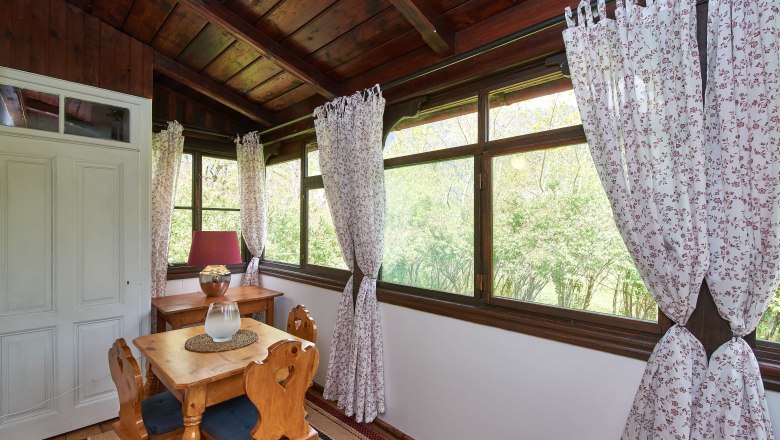Ein gemütlicher Wintergarten mit Holzmöbeln, floralen Vorhängen und Blick ins Grüne.