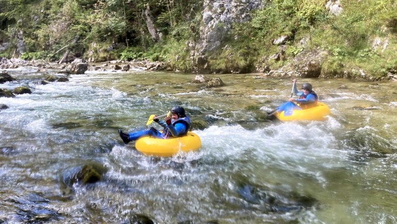 Zwei Personen beim Tubing auf einem Fluss in einer bewaldeten Umgebung.
