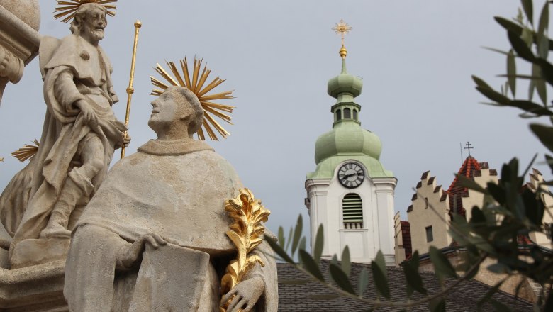 Statue mit Heiligenschein vor Kirchturm mit Uhr und gr&uuml;nem Dach.