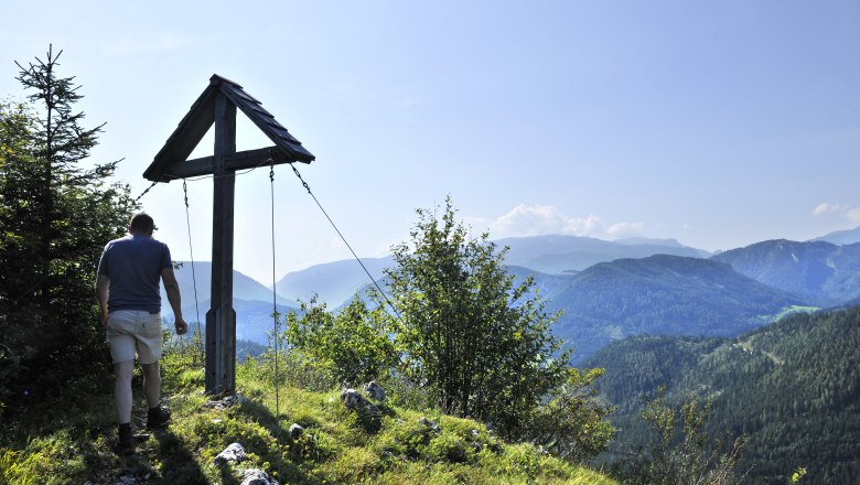 Ein Wanderer steht neben einem Gipfelkreuz mit Blick auf bewaldete Berge.