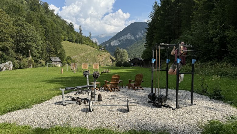 Outdoor-Fitnessbereich mit Trainingsger&auml;ten in einer gr&uuml;nen, bergigen Landschaft.