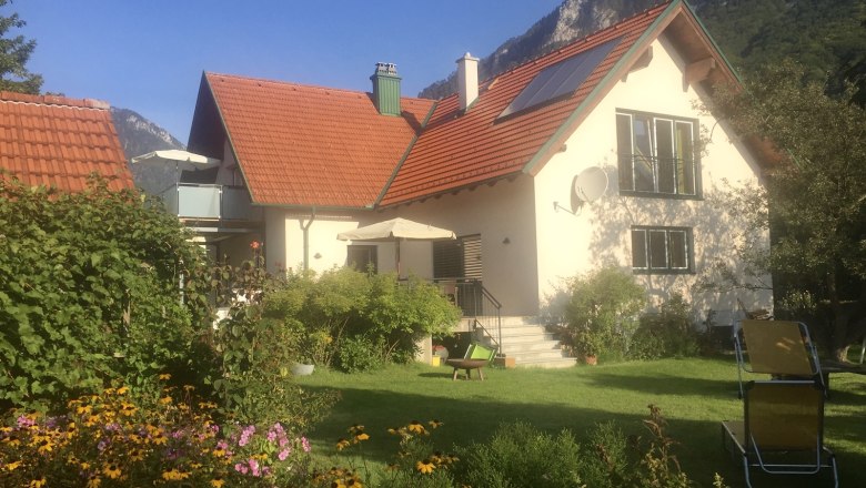 Ein Haus mit rotem Dach, umgeben von einem Garten mit Blumen und Liegestühlen, vor einer Bergkulisse.