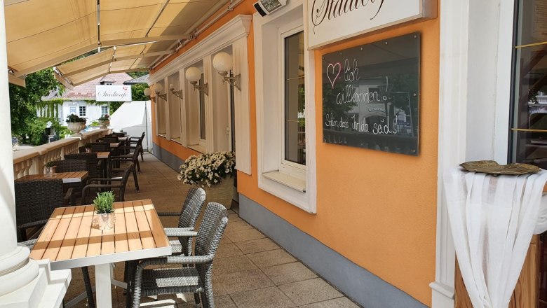 Außenbereich eines Cafés mit Tischen und Stühlen, orangefarbene Wand mit Schild 'Stadtcafé'.