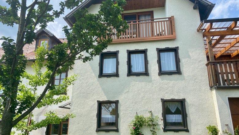 Ein traditionelles Haus mit weißen Wänden, Holzfenstern und einem Balkon. Ein Baum und Pflanzen im Vordergrund.