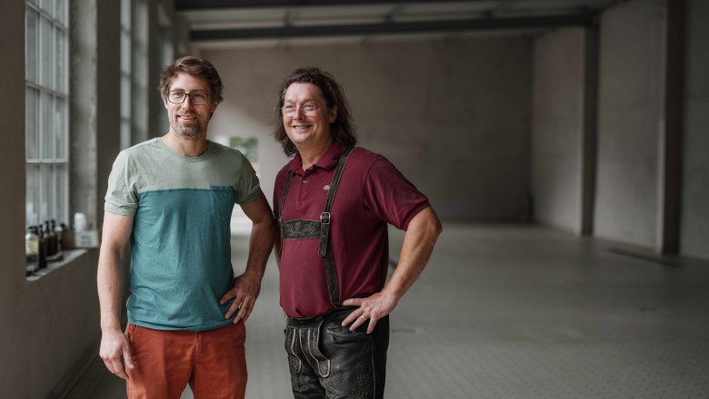 Zwei Männer, die Gründer von Raxbräu, stehen in einem leeren Raum, einer trägt ein T-Shirt und der andere Lederhosen.