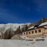 Ein Holzhaus im Schnee vor einer Bergkulisse unter klarem, blauem Himmel.