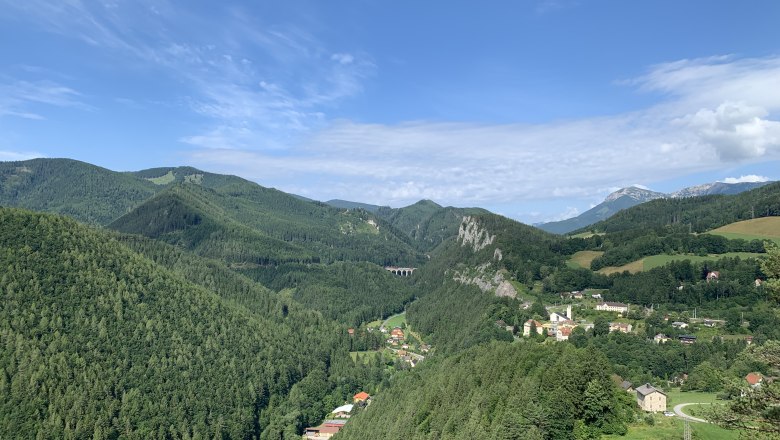 Panoramablick auf eine grüne Berglandschaft mit einem kleinen Dorf und einem Viadukt.