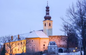 Schloss Gloggnitz im Winter mit beleuchtetem Turm und verschneitem Dach.