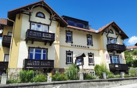 Gelbes Gebäude mit der Aufschrift 'WIENERHEIM', Holzbalkone und blauer Himmel.